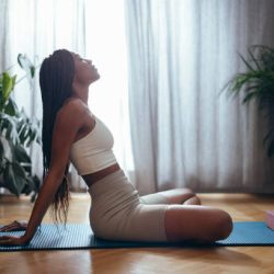Side view of young African-American woman sitting on blue yoga mat ready for workout session or yoga training with foam blocks at home